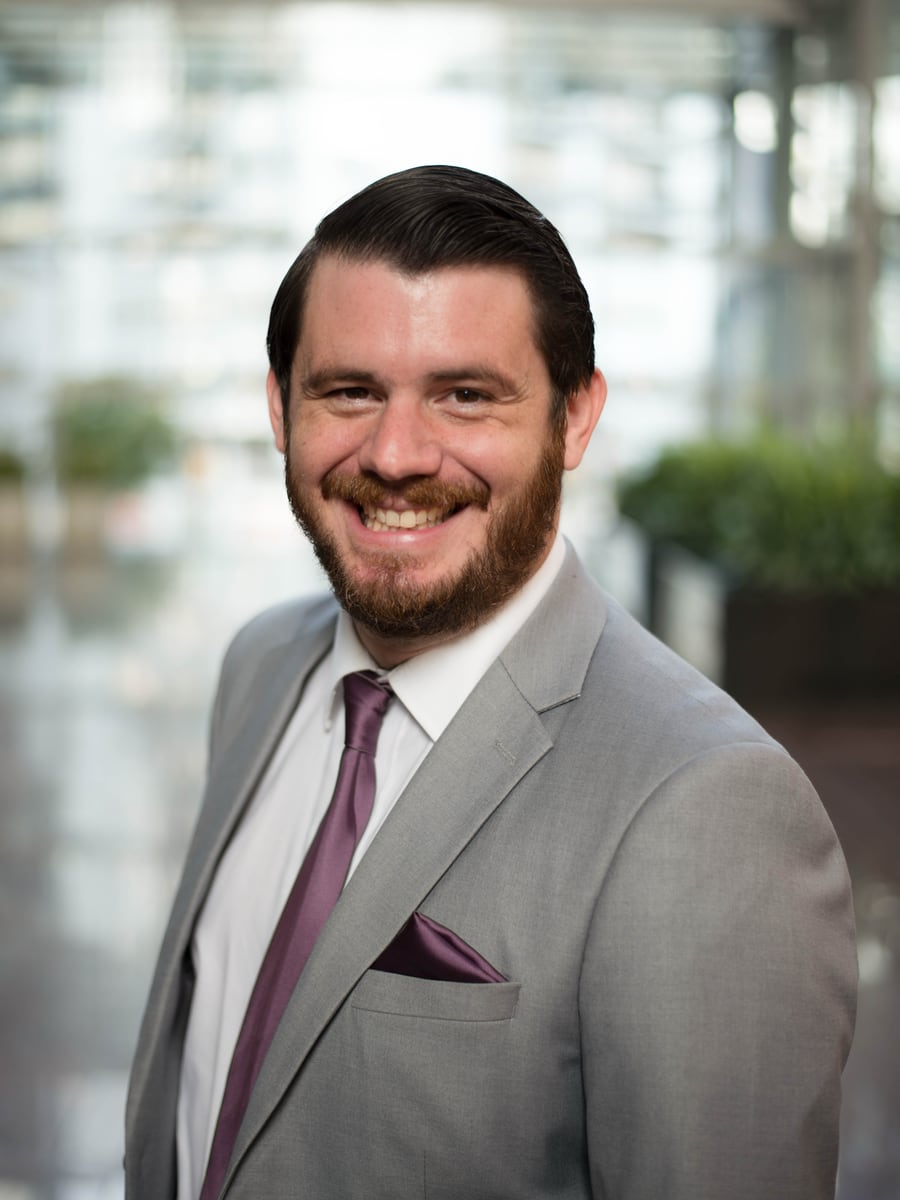 Zak Lyons — gray suit, plum tie, a wide smile, in a bright glass atrium. The professional headshot.